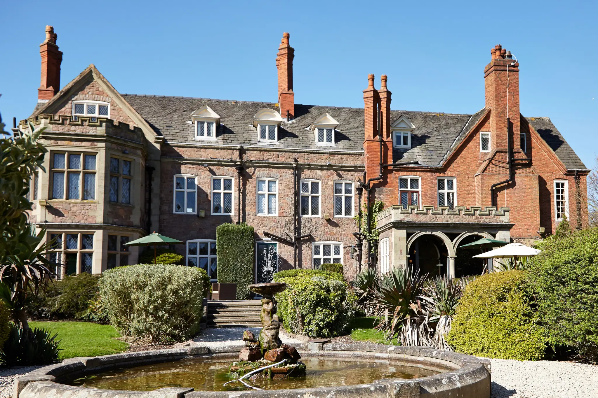 A historic brick mansion with numerous chimneys, a garden, and a stone fountain in the foreground under a clear sky. - Home Instead