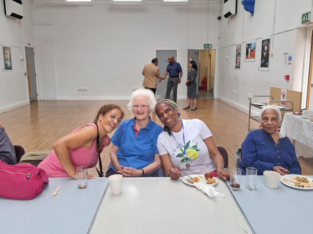 Three women sitting at a table, smiling, with a mostly empty hall in the background. - Home Instead