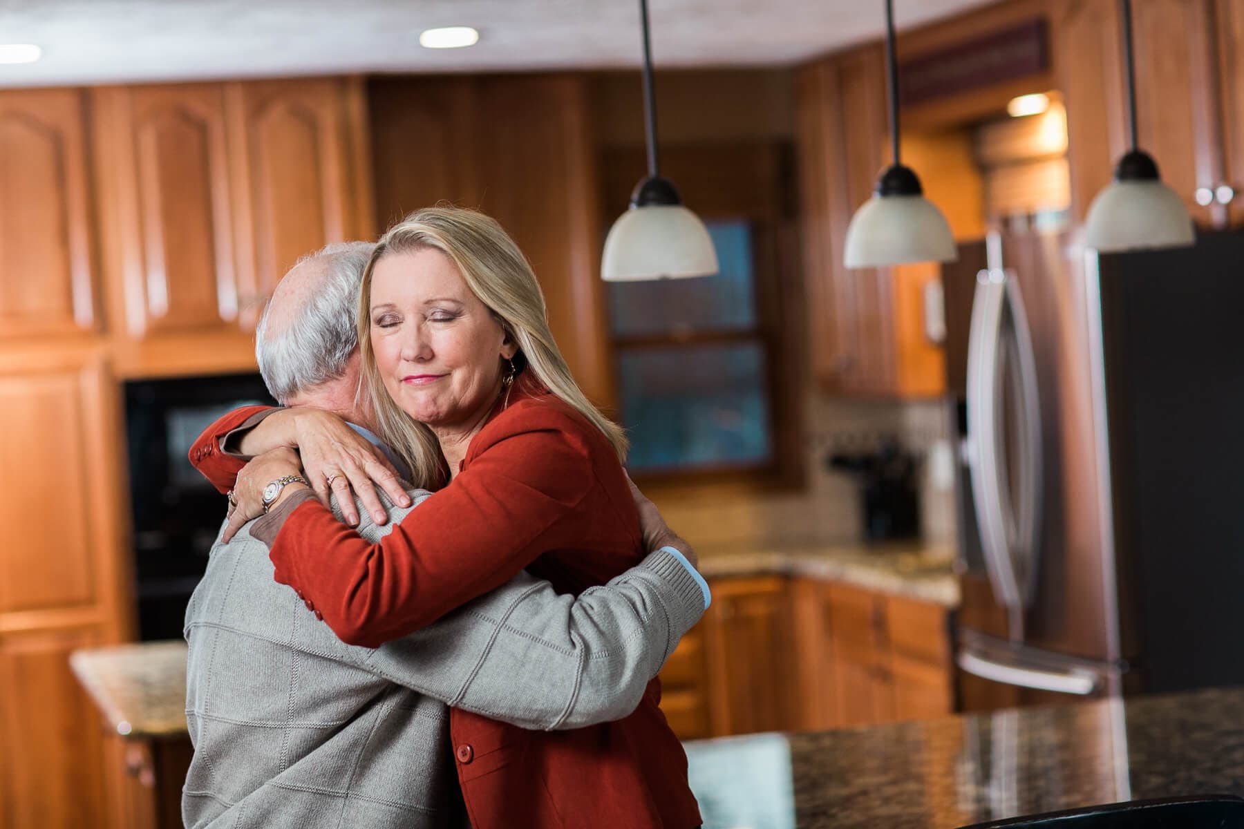 A man and woman hug in a warmly lit kitchen, with wooden cabinets and hanging lights in the background. - Home Instead