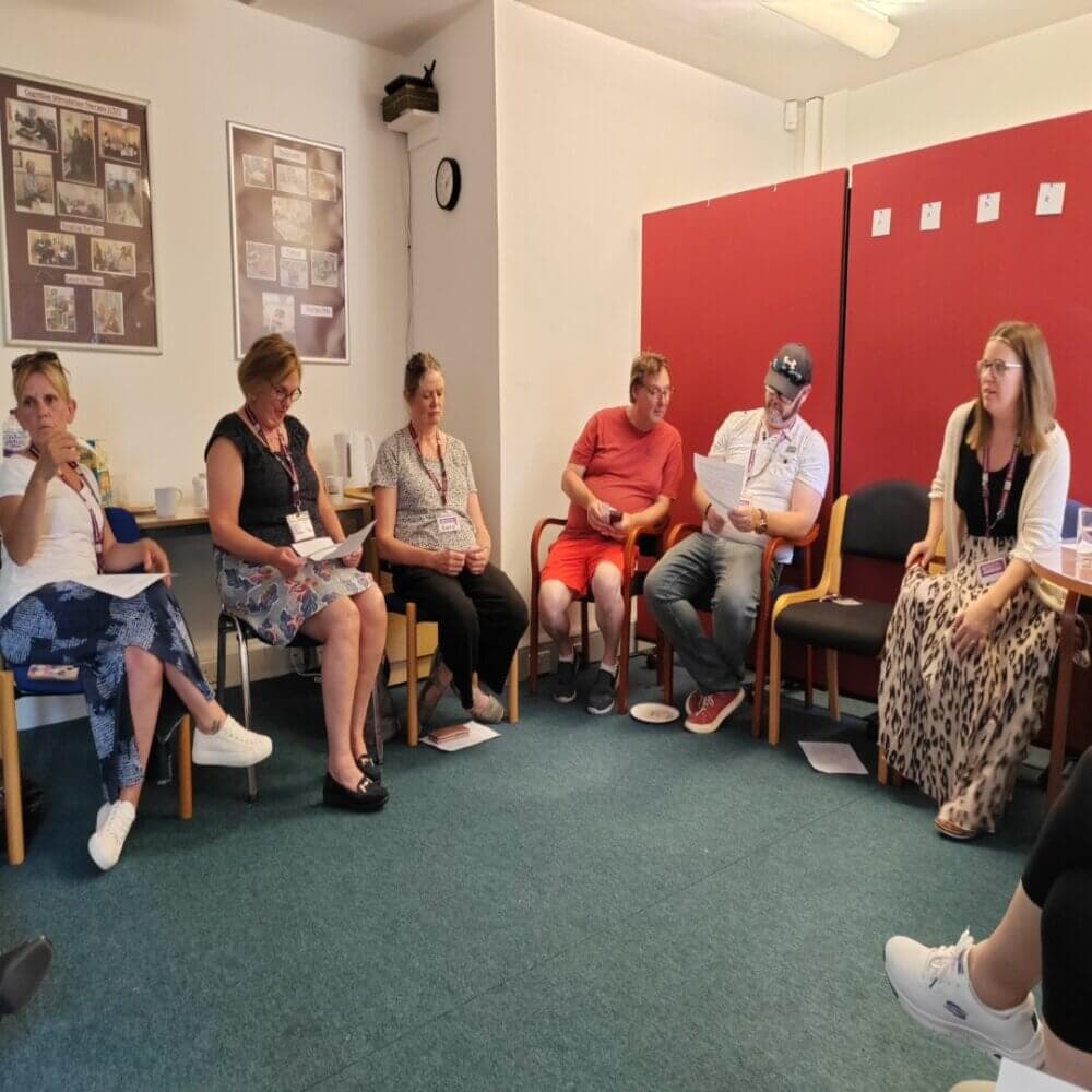 A group of seven people are sitting on chairs in a circle, engaging in a discussion in a room with red walls and posters. - Home Instead