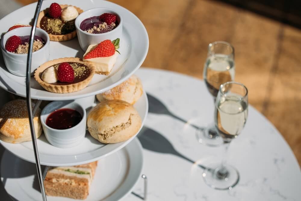 A tiered tray of pastries, scones, and sandwiches, accompanied by two glasses of champagne on a white table. - Home Instead