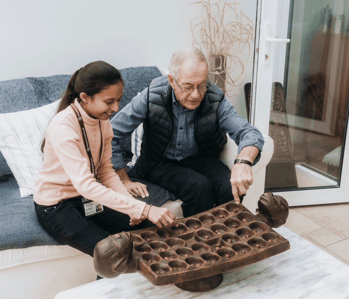 An elderly man and a young girl playing a traditional mancala game on a carved wooden board while sitting on a couch. - Home Instead