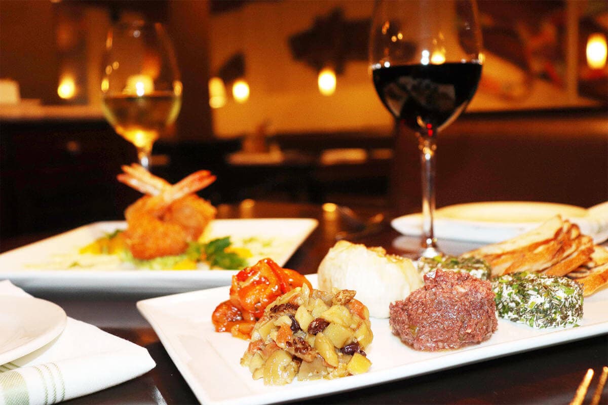 Close-up of a table with white plates of assorted appetizers, shrimp, and two glasses of red and white wine. - Home Instead
