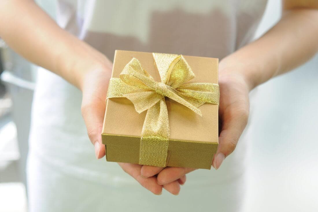 Person holding a gold gift box with a shiny ribbon bow. - Home Instead