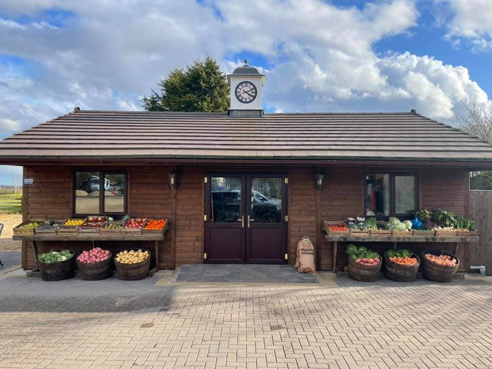 Wooden farm store with clock tower, displaying a variety of fresh vegetables in barrels out front on a sunny day. - Home Instead