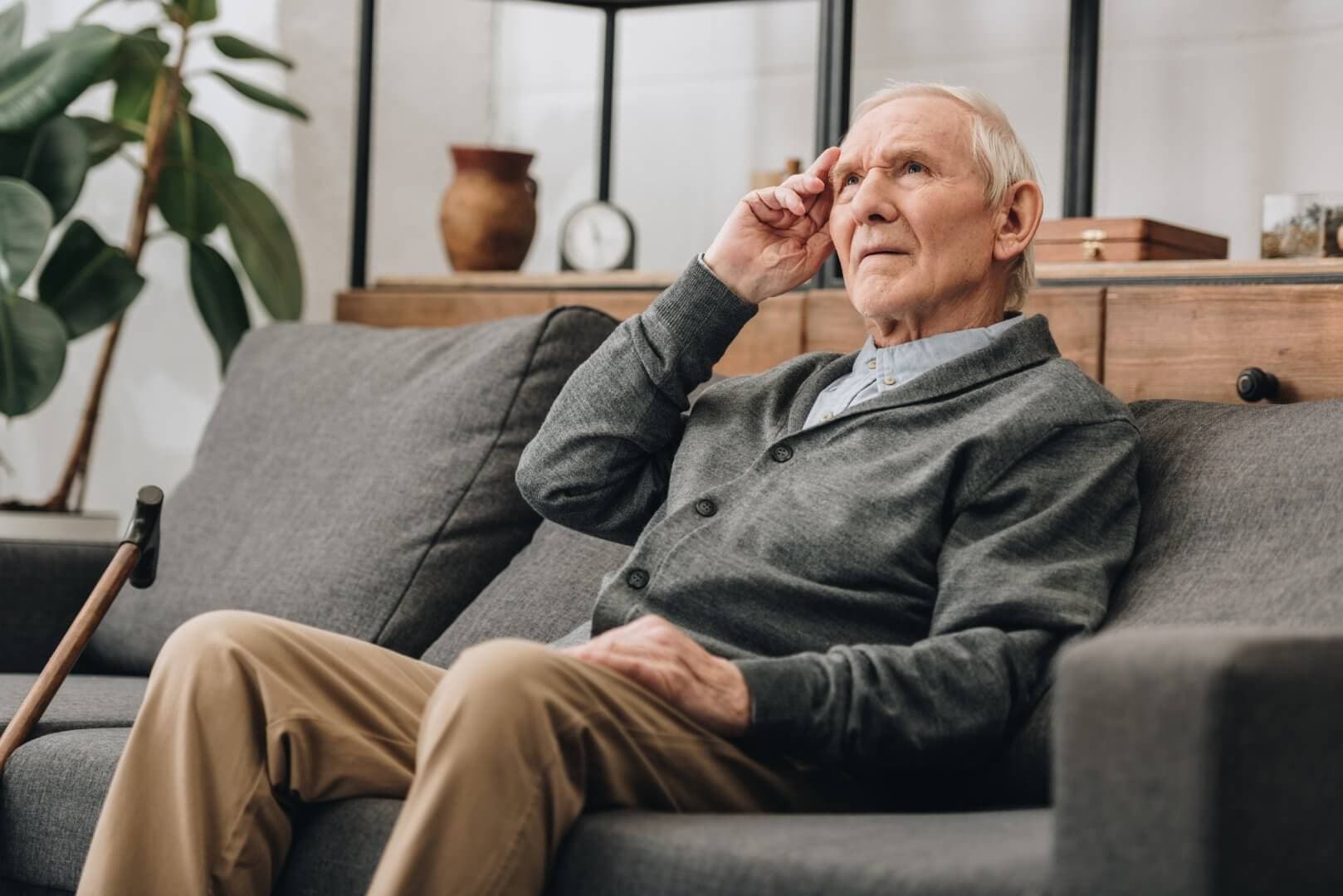 Elderly man in a grey cardigan sitting on a sofa, touching his temple, with a cane nearby and a plant in the background. - Home Instead