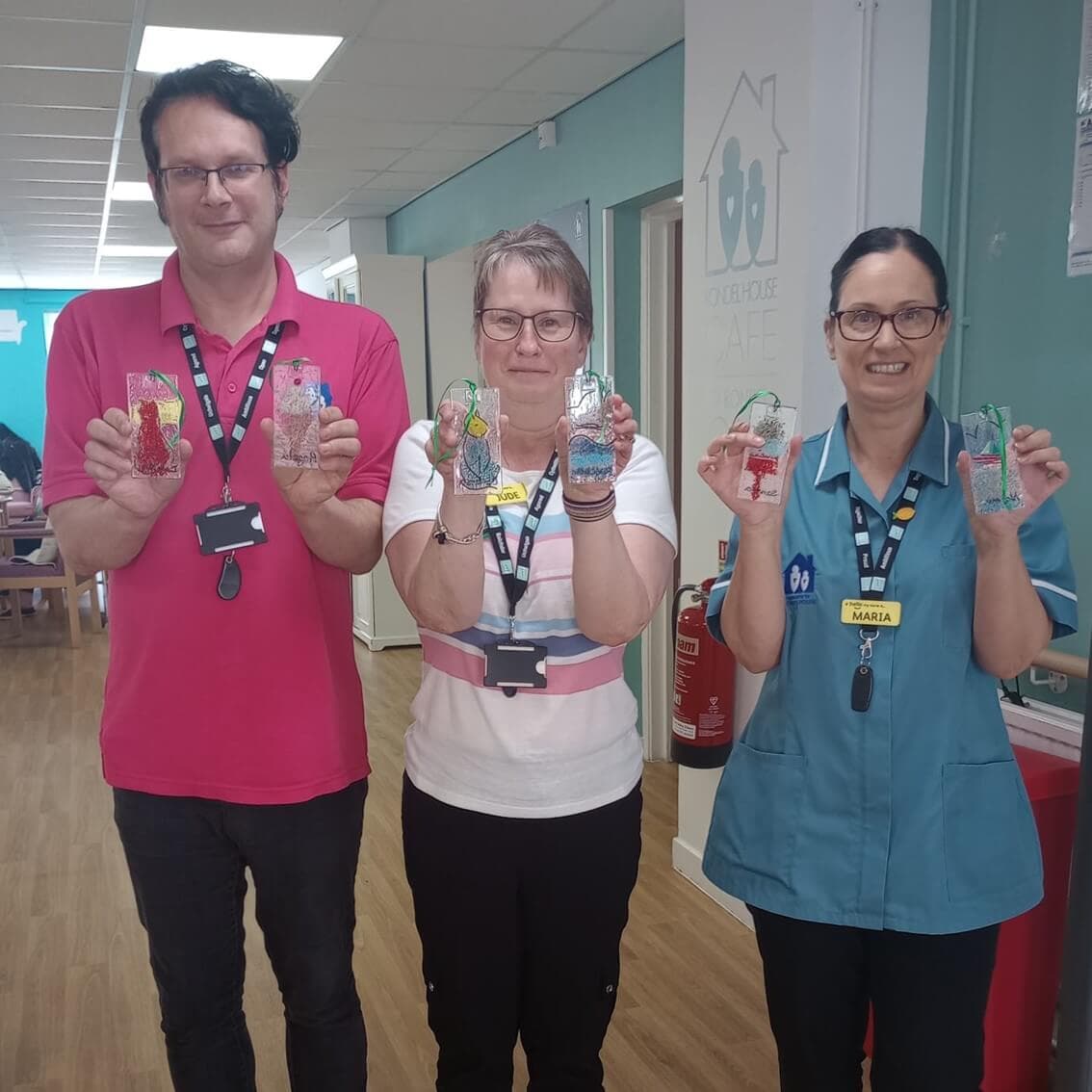 Three people standing indoors, holding up decorated jars, and wearing badges around their necks. - Home Instead
