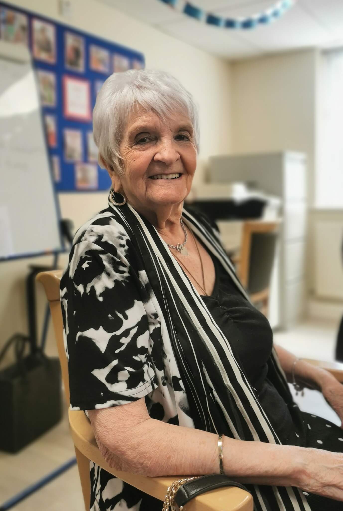 Elderly woman with short white hair smiling, seated indoors, wearing a black and white outfit and pendant necklace. - Home Instead