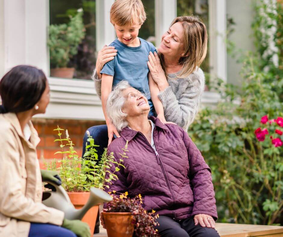 A elderly woman sitting and smiling at a child, with two adults nearby in a garden. - Home Instead