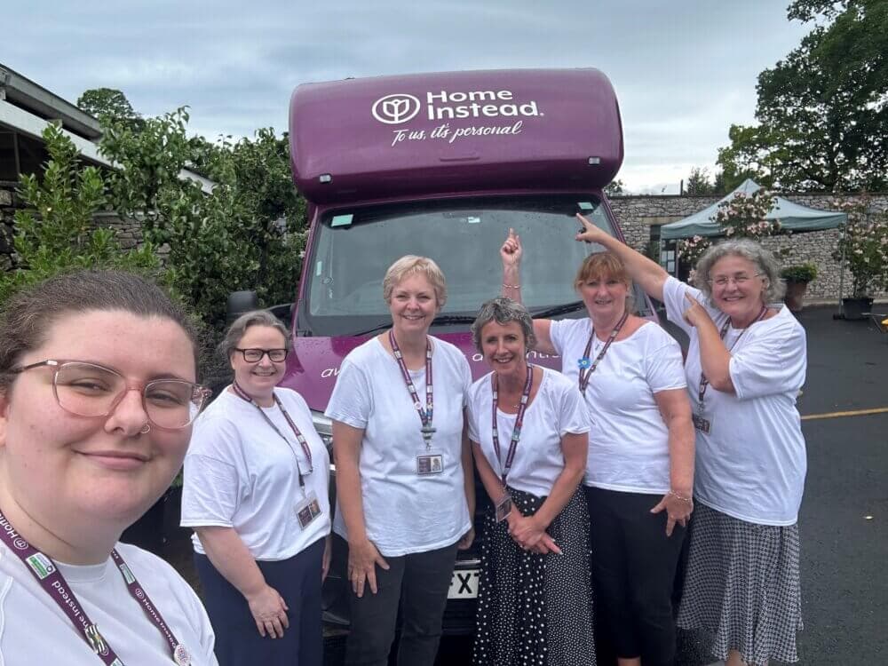 Group of six women wearing white shirts standing in front of a purple Home Instead van, smiling and posing. - Home Instead