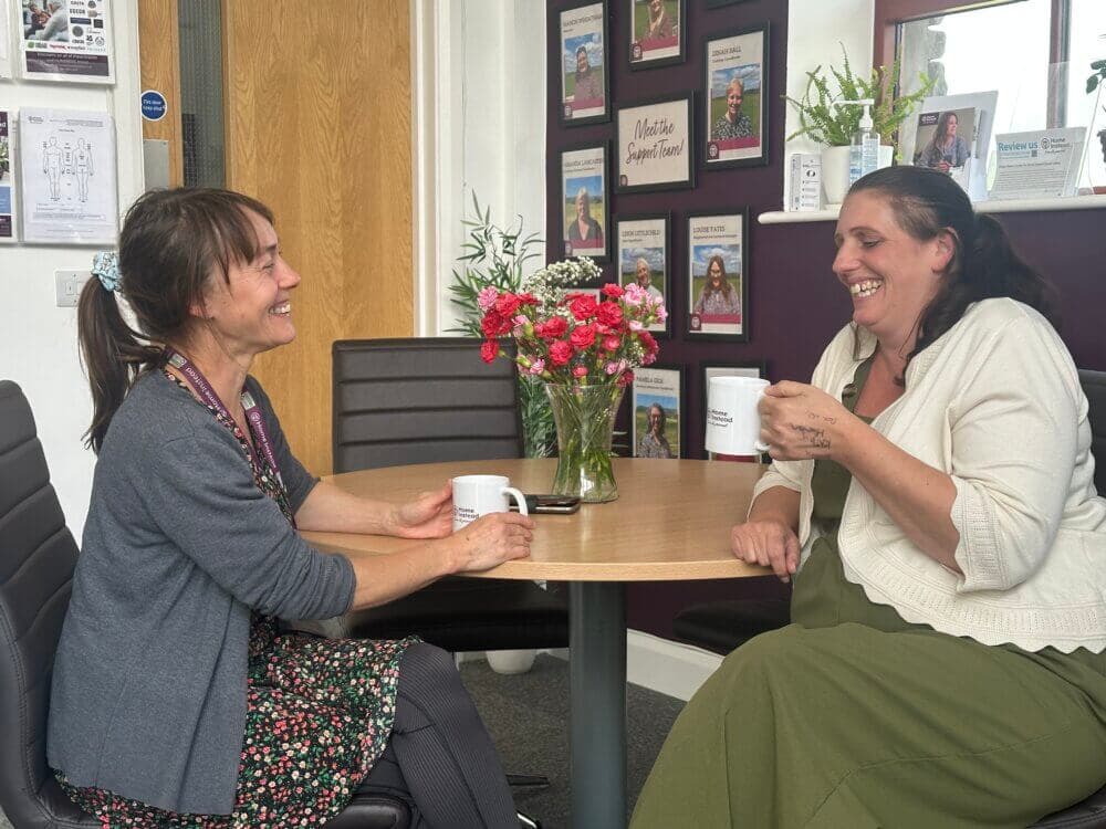 Two women smiling and having coffee together at a small table in an office setting, with a vase of flowers in the middle. - Home Instead