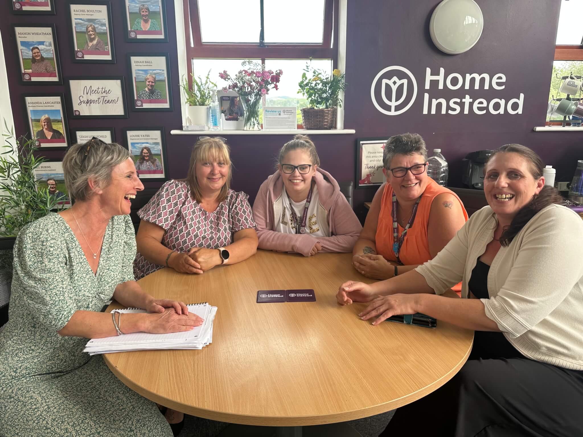 Five women sitting around a table, smiling and discussing in an office with awards on the wall and "Home Instead" logo. - Home Instead