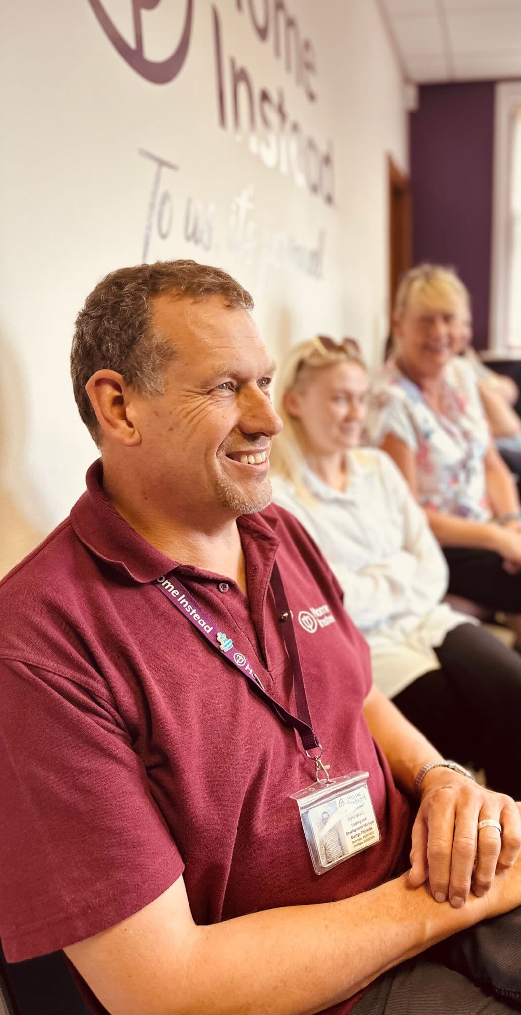 Smiling person in a maroon shirt and badge sits with others in a brightly lit room, text "Home Instead" on the wall behind. - Home Instead