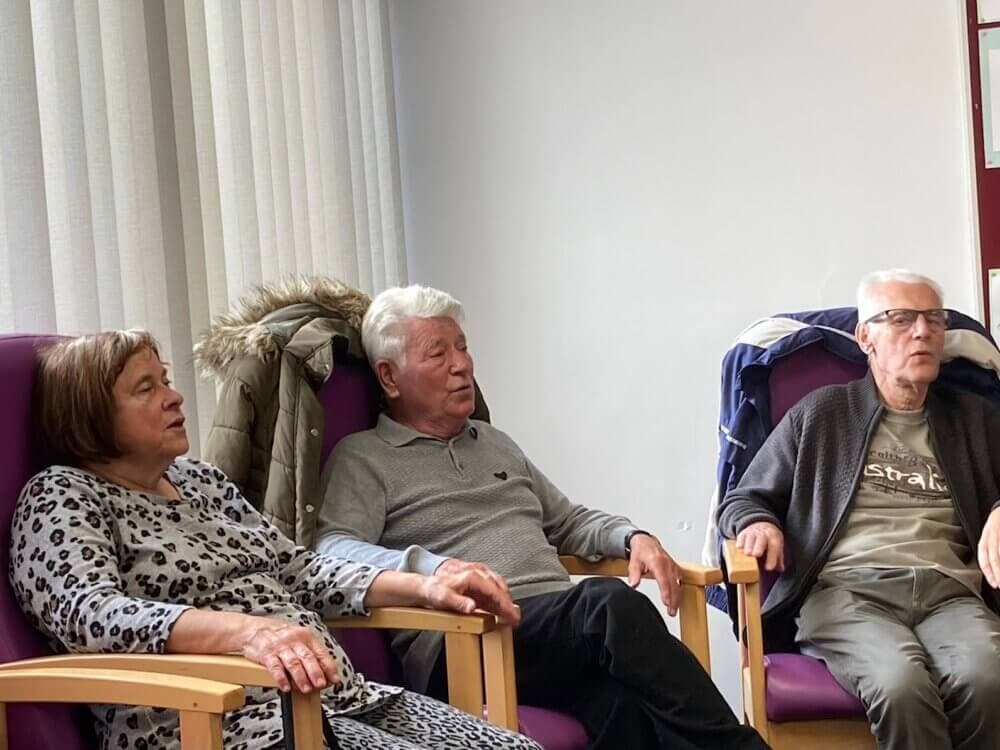 Three elderly people are sitting in chairs, with coats draped over the chairs behind them. - Home Instead