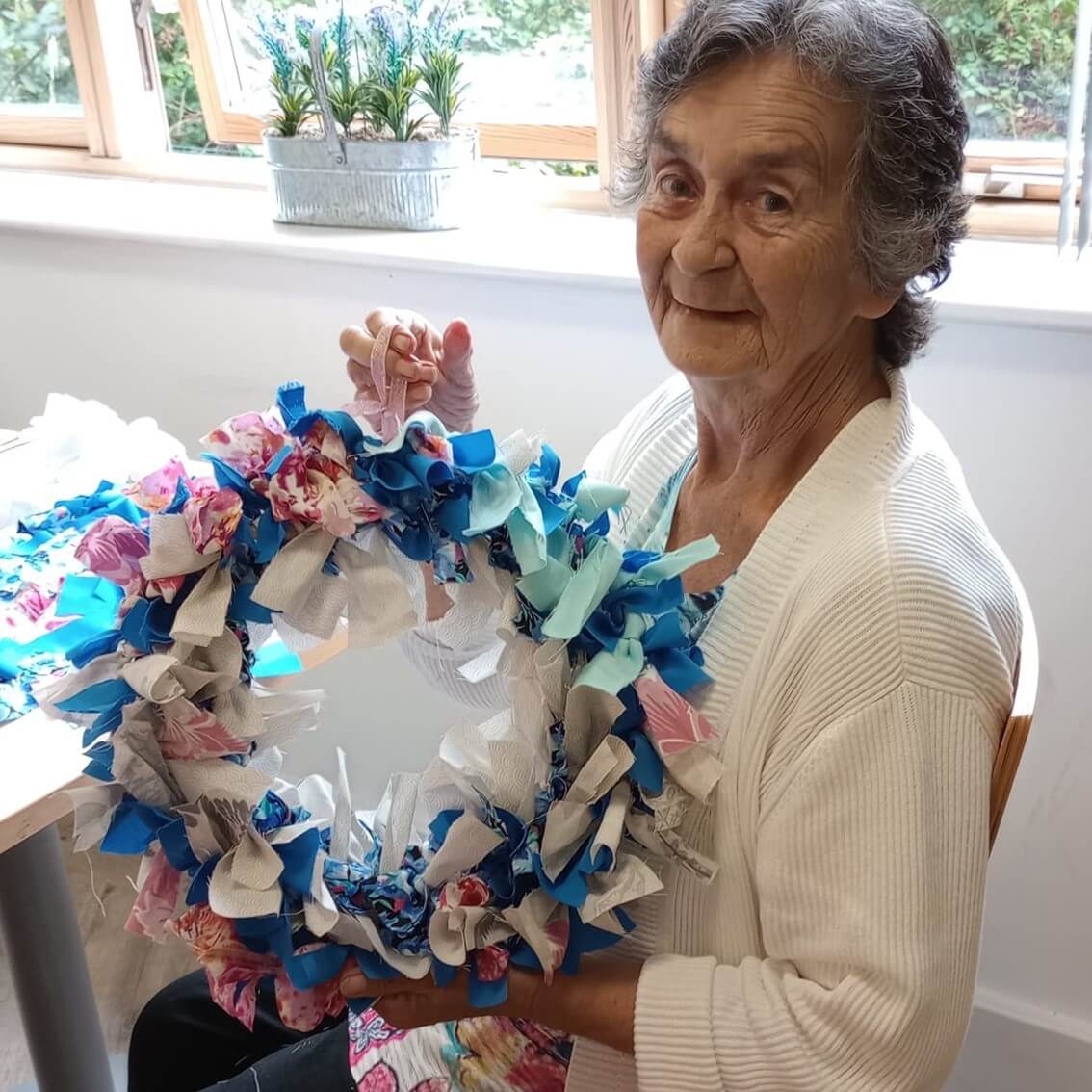An elderly woman smiles while holding a colorful handmade wreath with blue, white, and pink fabric. - Home Instead