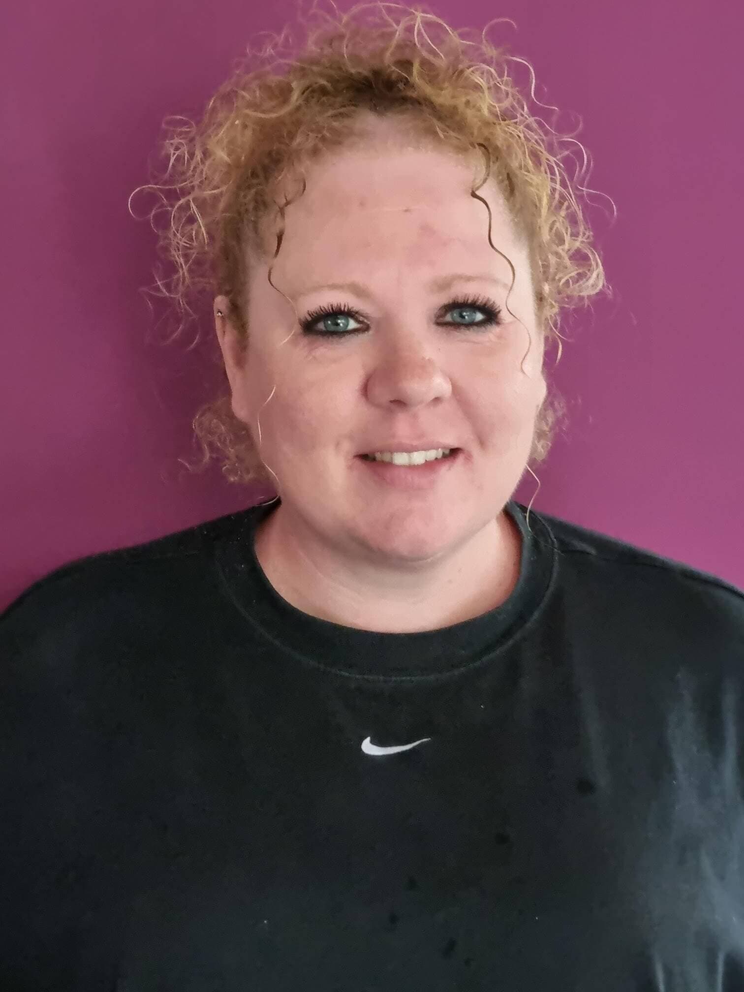 Smiling person with curly hair against a purple background, wearing a black Nike shirt. - Home Instead