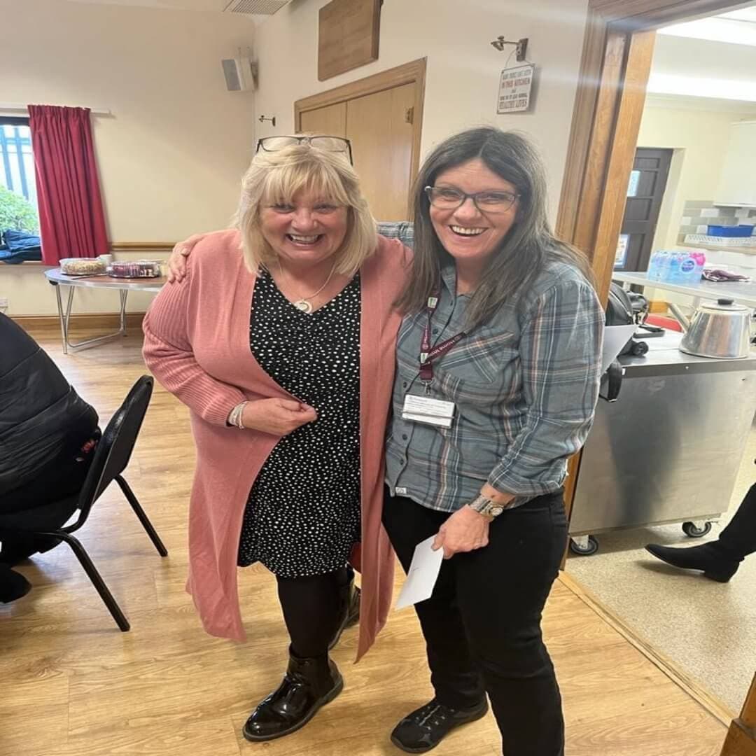 Two women smiling together in a room, one in a pink cardigan and dress, the other in a plaid shirt and badge. - Home Instead