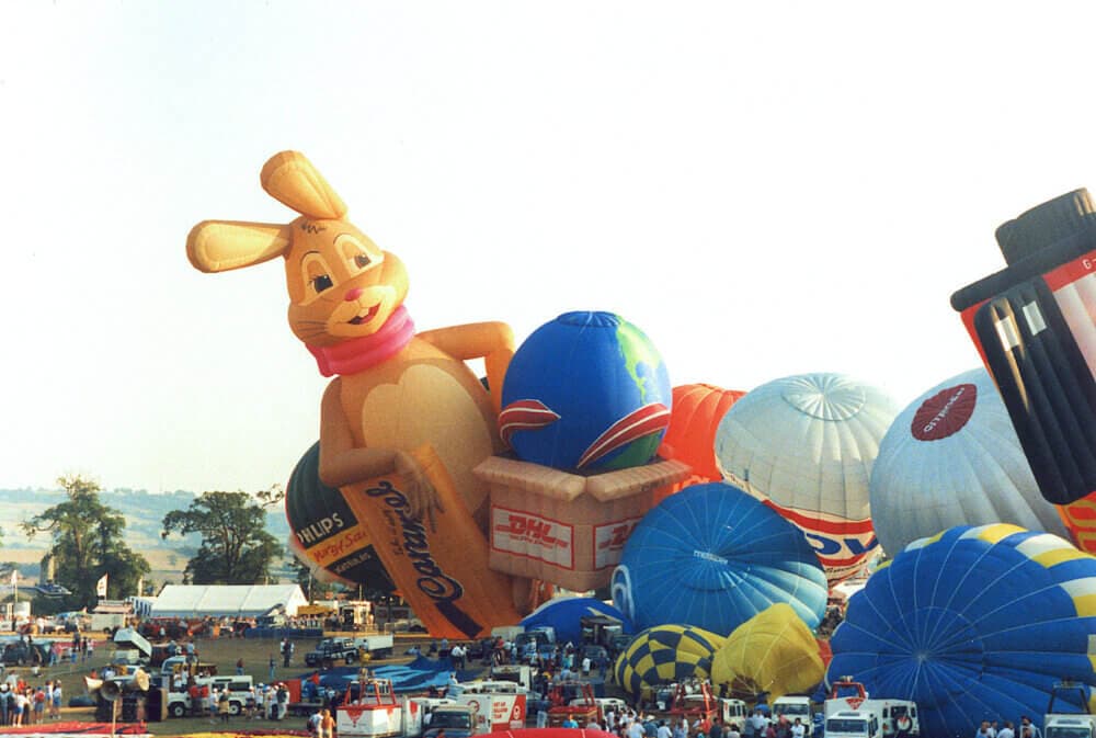 Hot air balloons with various designs, including a bunny and a basket, at a festival with many people and vehicles around. - Home Instead