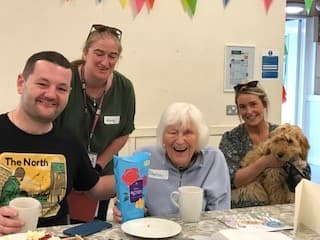 Four cheerful people sitting at a table with a dog, enjoying snacks and drinks indoors, bunting in the background. - Home Instead