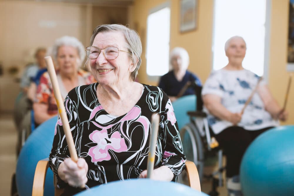 Elderly woman smiling and participating in a group drumming exercise with others in a bright room. - Home Instead
