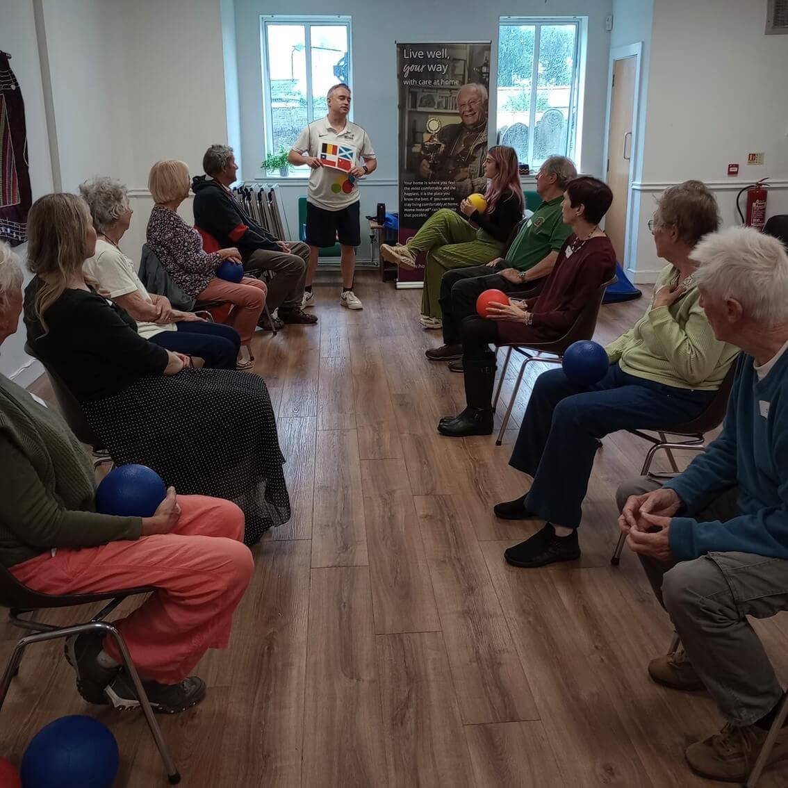 A group of seniors sit in a circle with exercise balls while a leader stands and talks to them in a bright room. - Home Instead