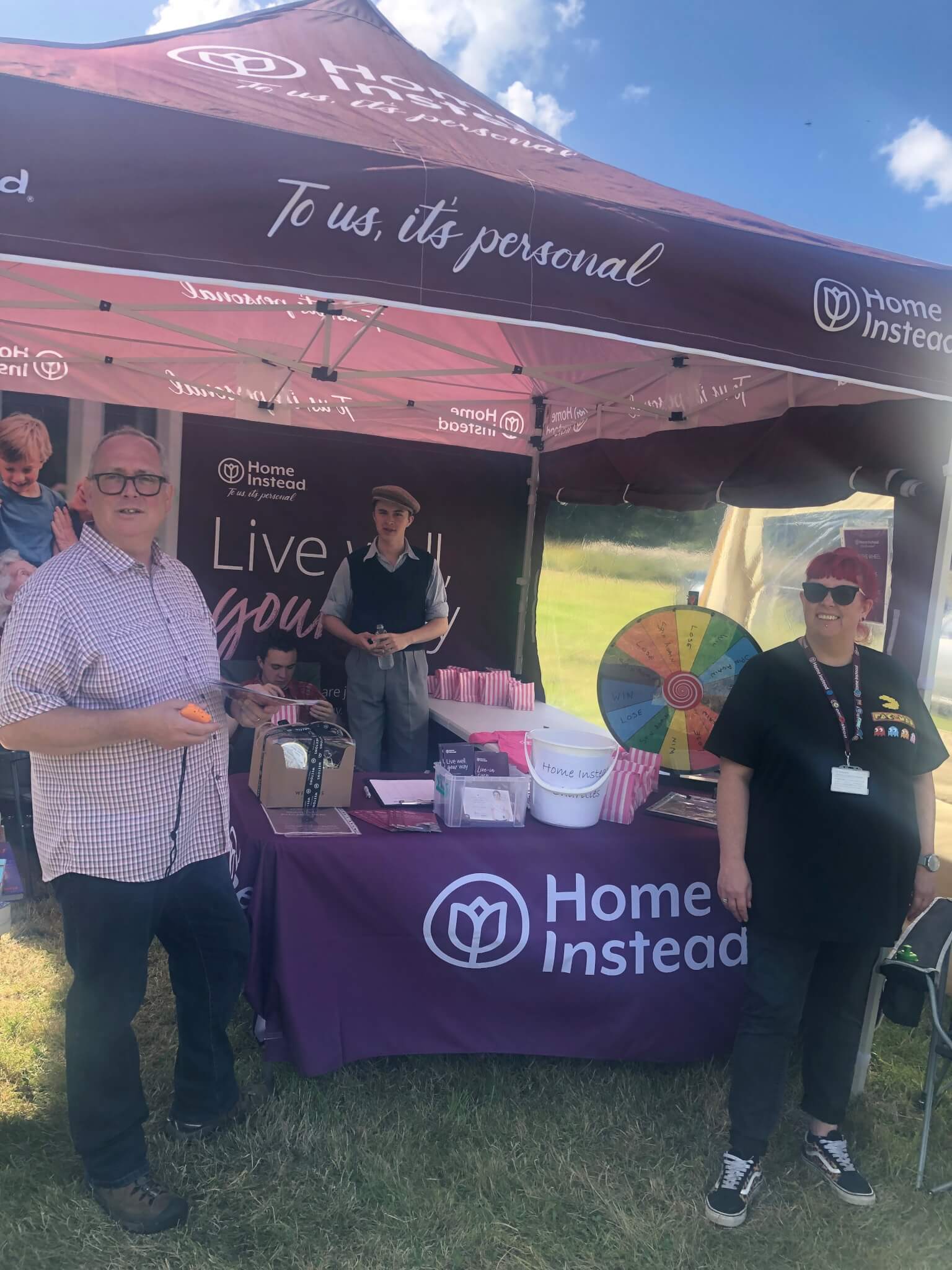 People standing under a Home Instead tent with a table, a spinning prize wheel, and promotional material on a sunny day. - Home Instead