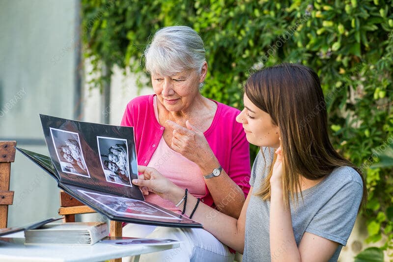 An older woman and a younger woman sitting outside, looking at a photo album together on a sunny day. - Home Instead