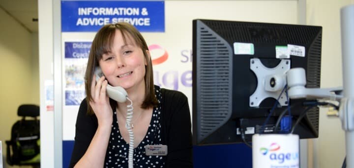 A woman is sitting at a desk, smiling and holding a phone to her ear, with an informational sign in the background. - Home Instead