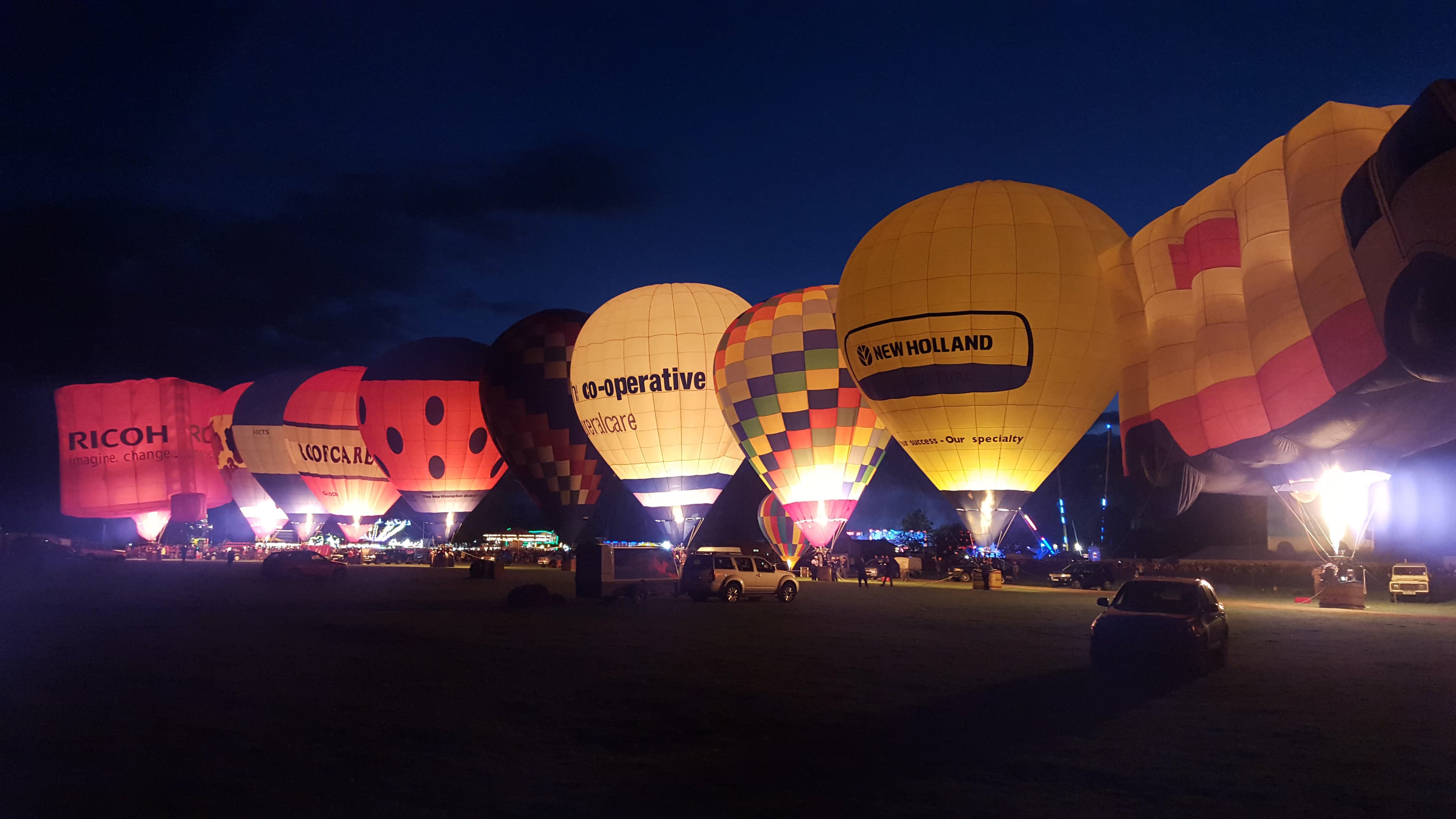 Hot air balloons light up at night, with various colorful patterns and logos, against a dark sky. - Home Instead