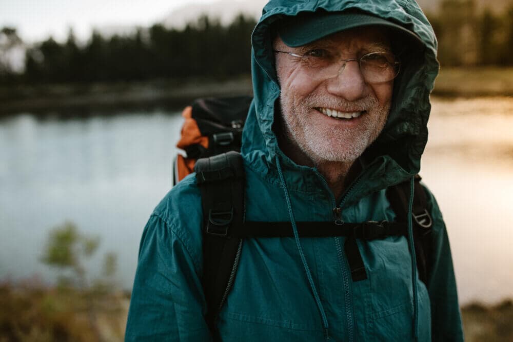 Elderly man with glasses and a hooded jacket smiling by a lake, carrying a backpack on a cloudy day. - Home Instead