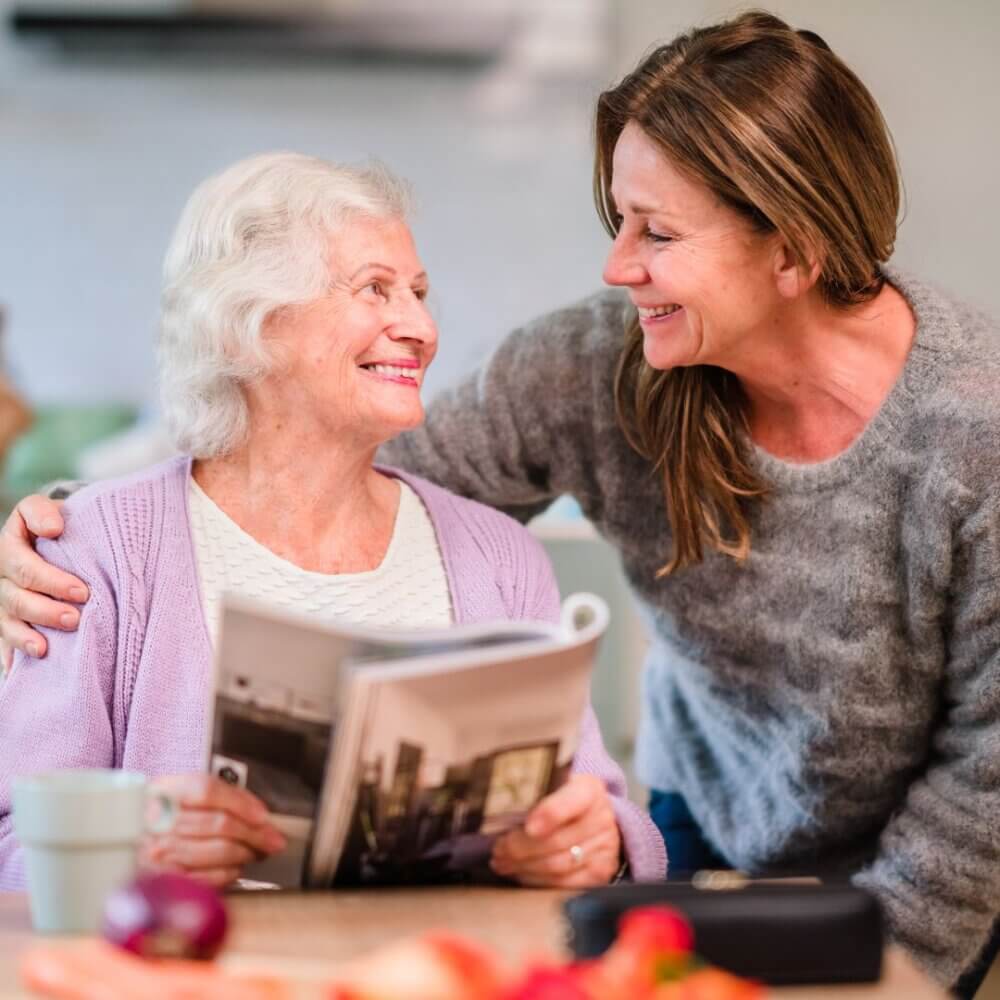 An elderly woman reading a magazine and a younger woman smiling at each other while hugging in a cozy setting. - Home Instead