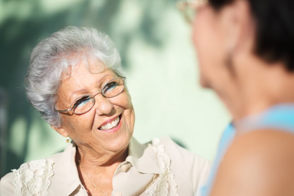 An elderly woman with glasses smiles at another person in the foreground, while sitting outside on a sunny day. - Home Instead