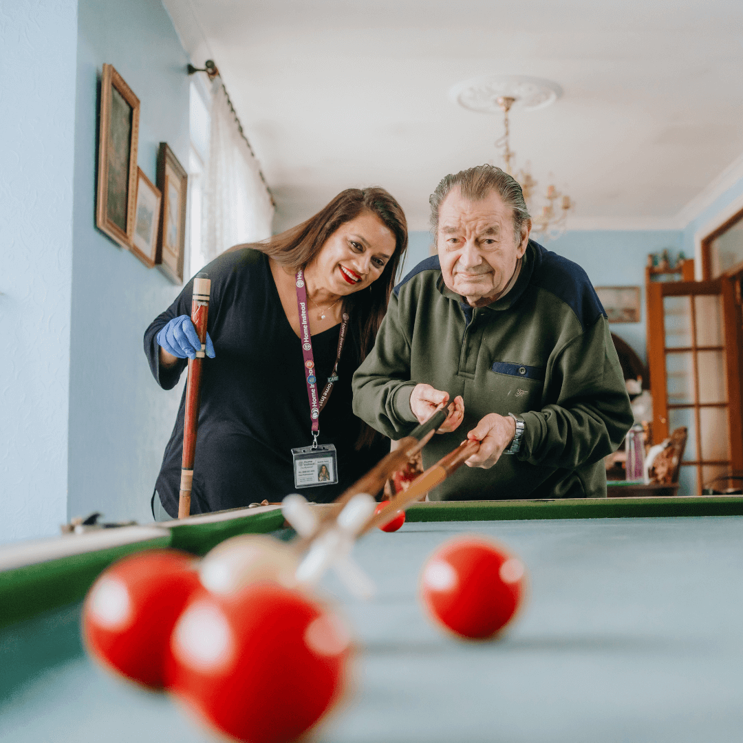 A woman assists an elderly man in playing a game of billiards in a well-lit room with blue walls. - Home Instead