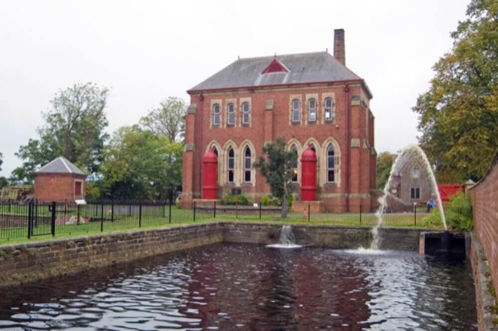 Red-brick historical building with arched windows by a canal, water fountain, and gated garden. - Home Instead