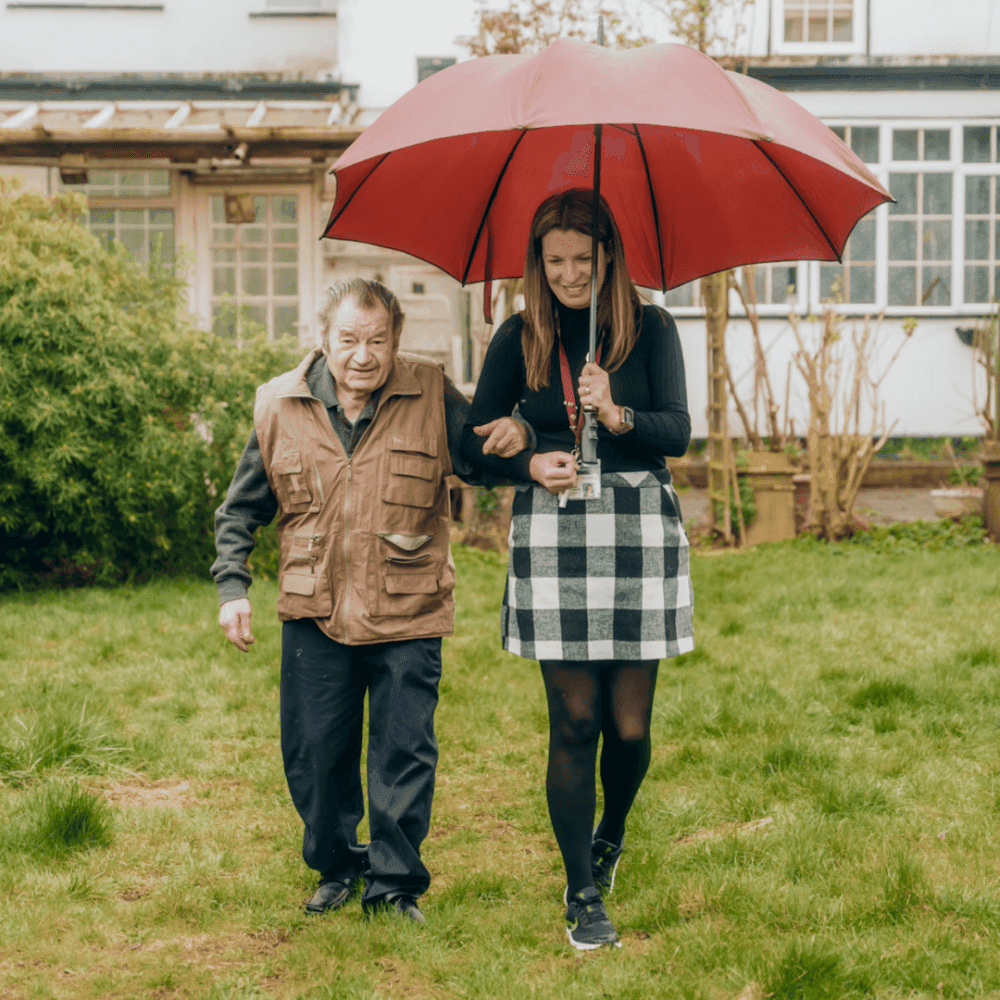 A young woman holding a red umbrella walks beside an elderly man, helping him outside in a garden. - Home Instead