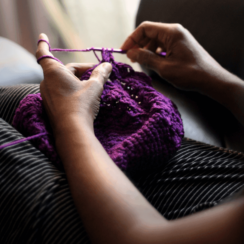 Close-up of a person knitting a purple item with needles, sitting on a striped surface. - Home Instead