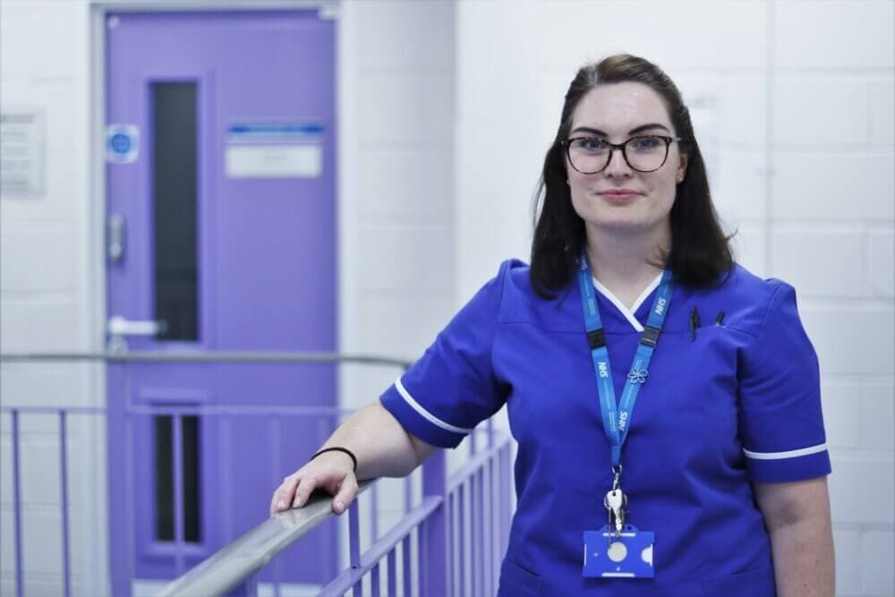 A woman in a blue nurse uniform stands in a hallway with purple doors and railings, smiling at the camera. - Home Instead