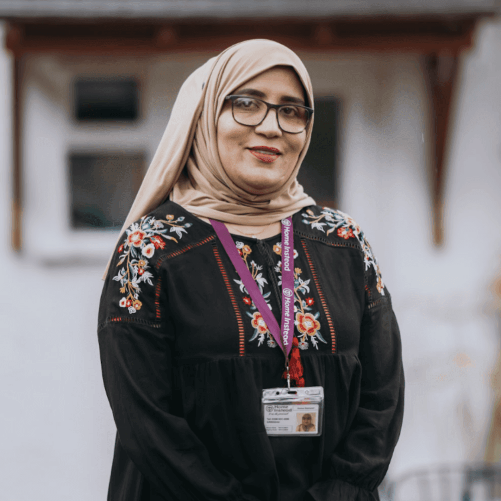 A woman wearing a hijab and glasses, dressed in a black embroidered top with a name tag around her neck, smiles at the camera. - Home Instead