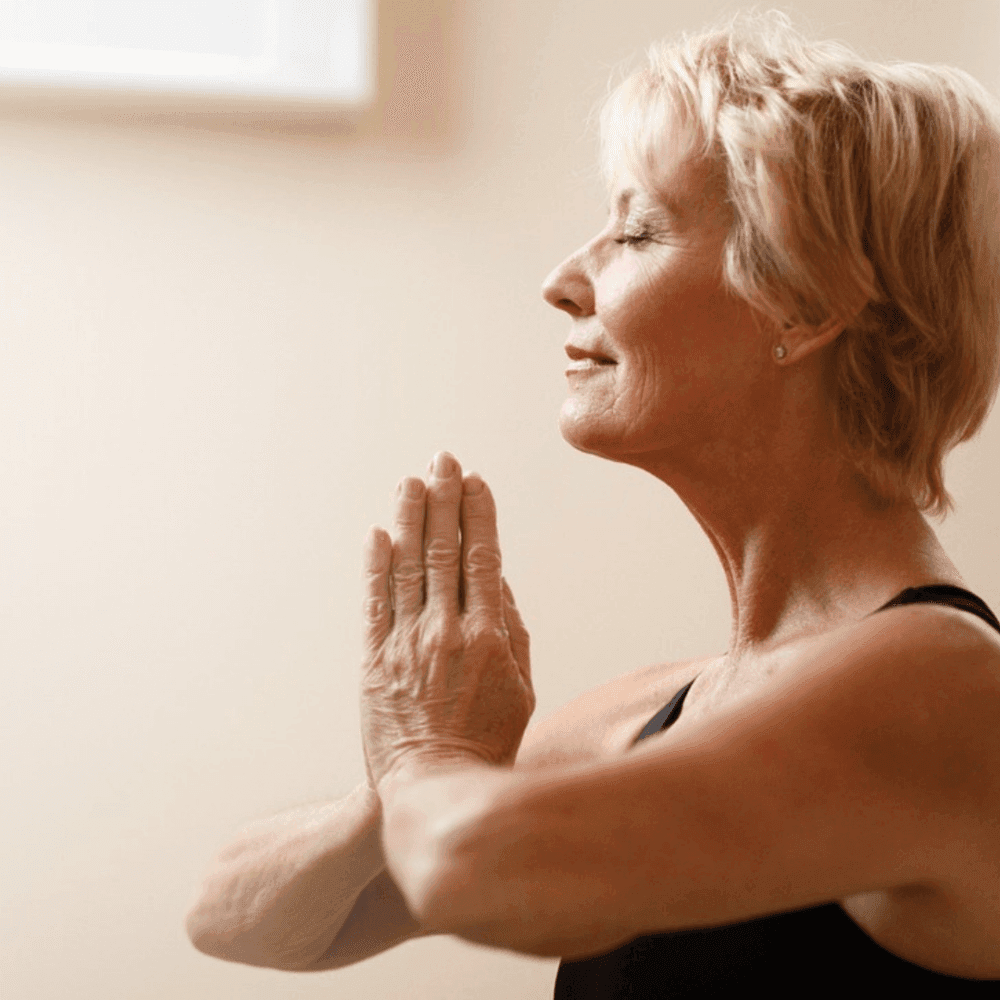 A person with short blond hair practices yoga, holding their hands in prayer position with eyes closed in a peaceful setting. - Home Instead
