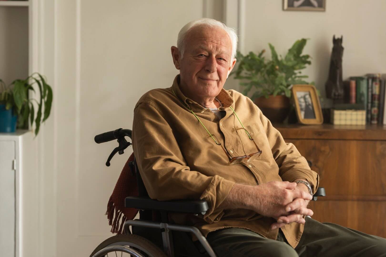 Older man sitting in a wheelchair, smiling, with hands folded on his lap, plants and bookshelf in the background. - Home Instead