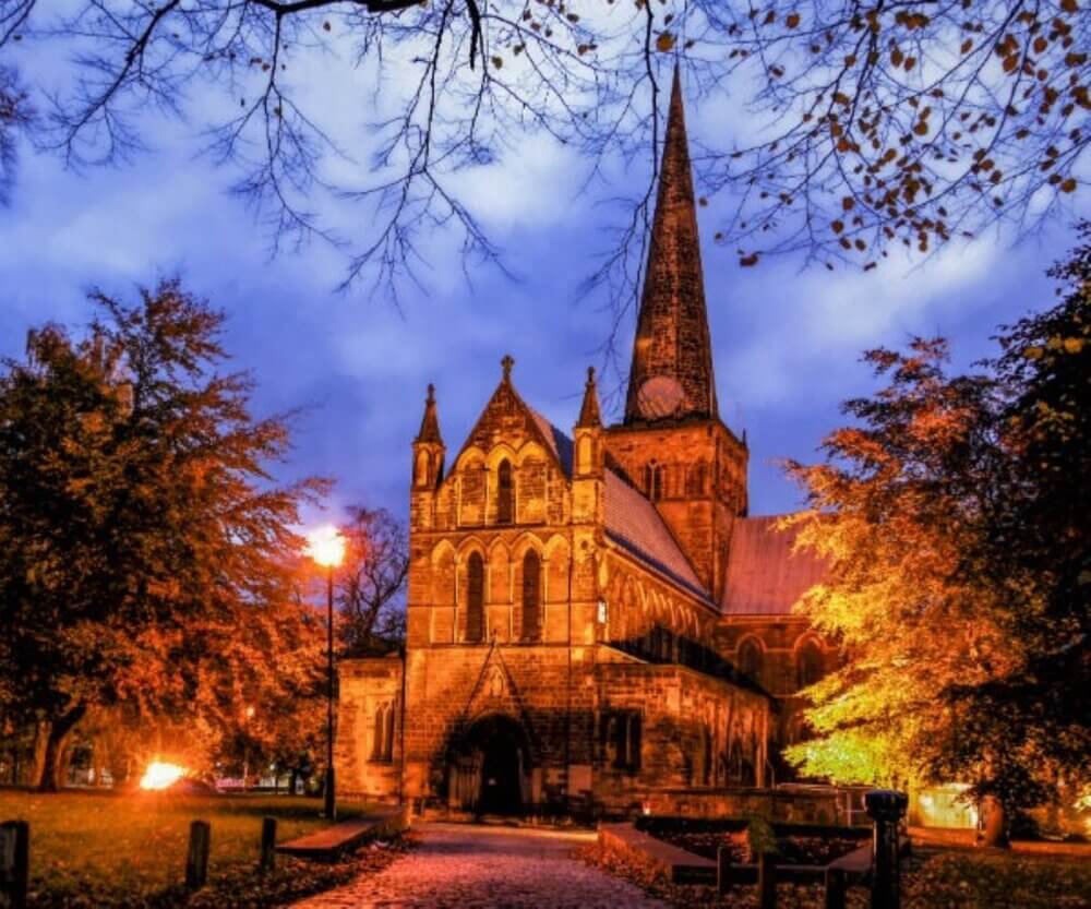 A historic church with a tall spire, surrounded by trees with autumn leaves, illuminated against a twilight sky. - Home Instead