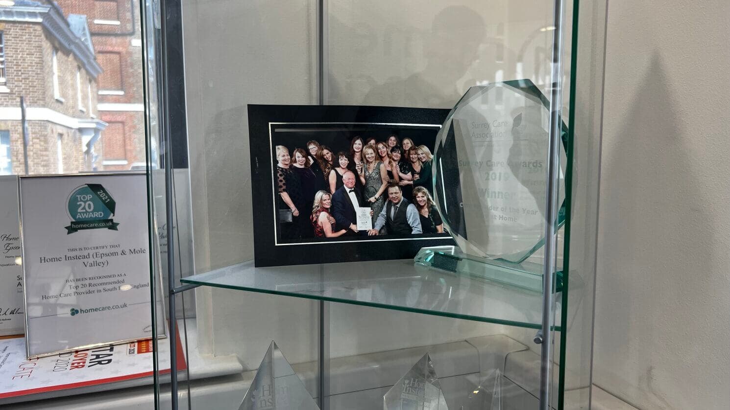 A framed group photo and an engraved award are displayed in a glass cabinet with other awards visible in the background. - Home Instead