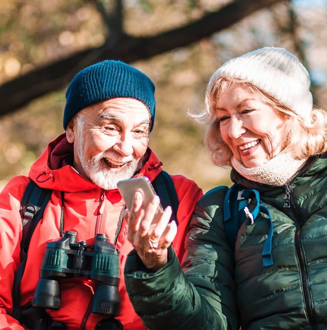 An elderly couple in outdoor gear smiles and looks at a smartphone screen together while sitting outside. - Home Instead