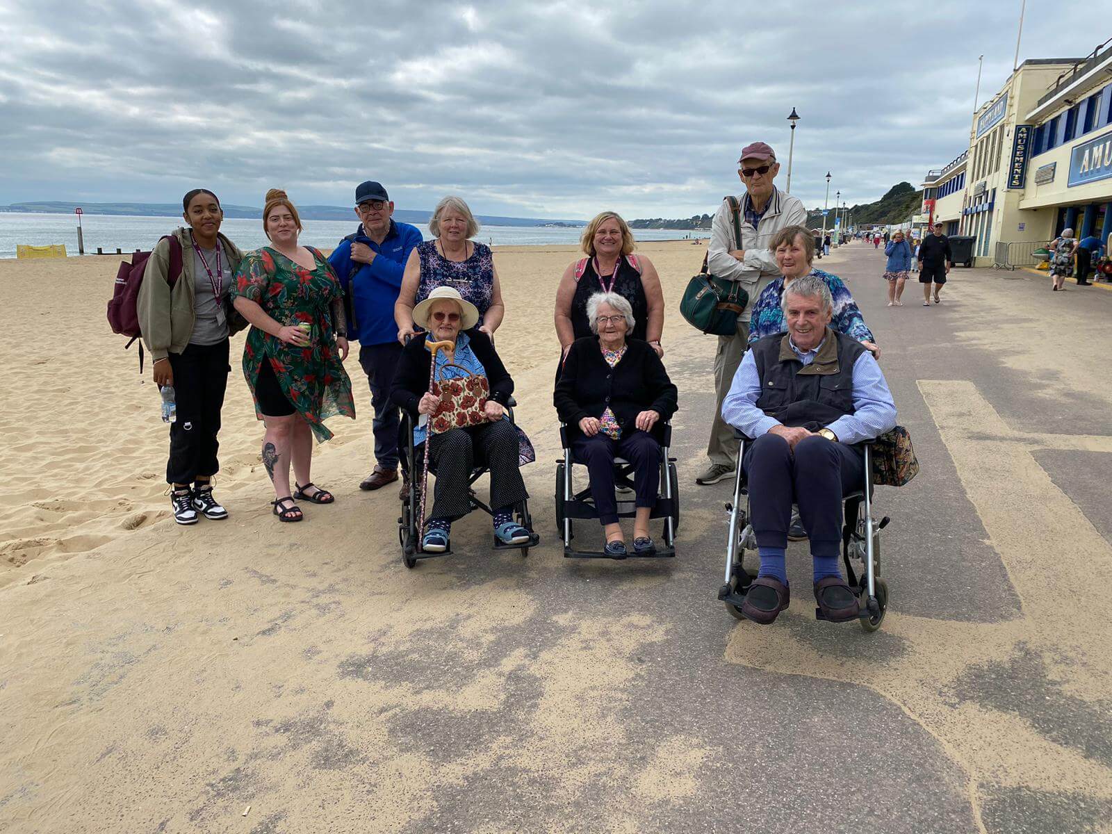 A group of people, including two in wheelchairs, smile on a beach promenade on a cloudy day. - Home Instead