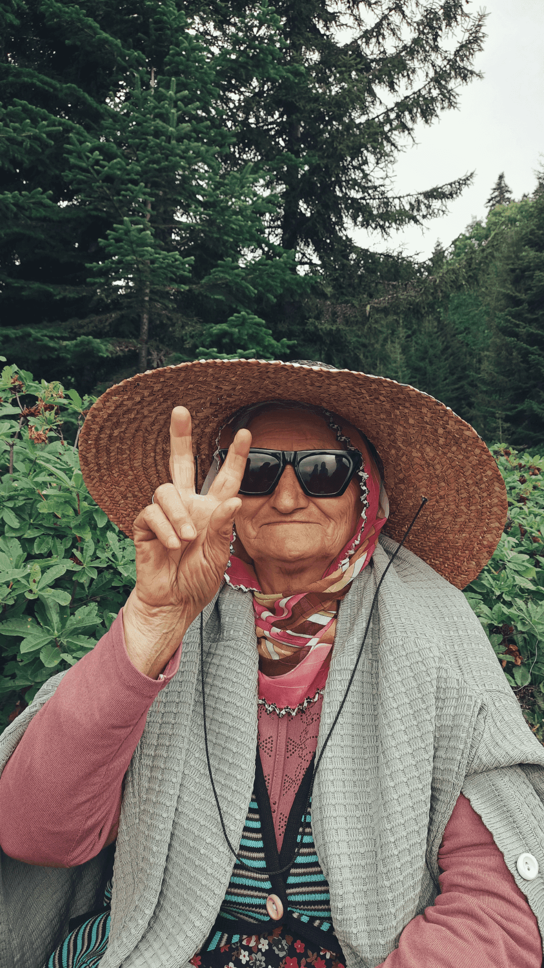 Elderly woman in a straw hat and sunglasses flashes a peace sign, standing in front of a forest background. - Home Instead