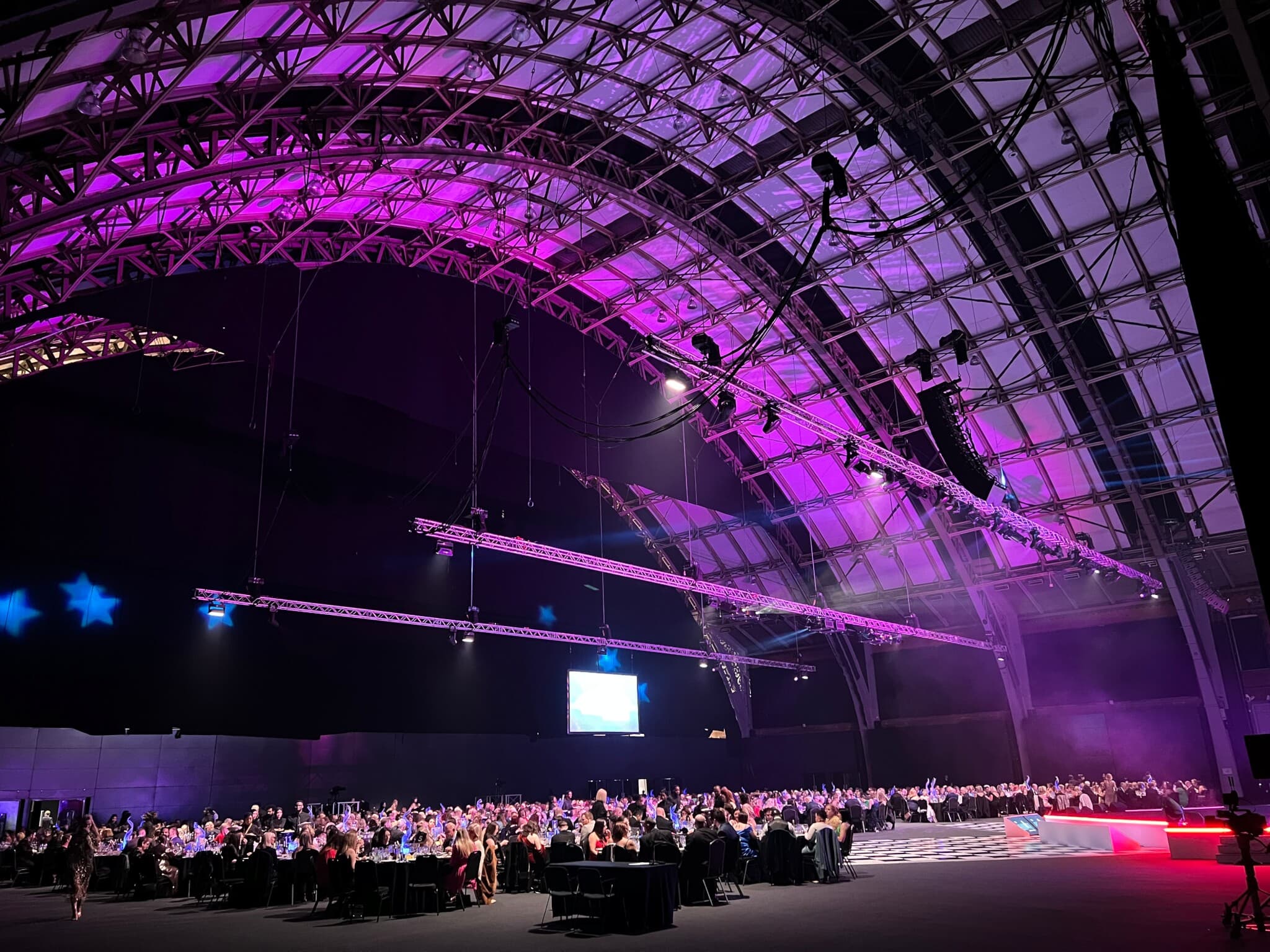 Large event in a spacious venue with high arched ceilings, purple lighting, and guests seated at round tables. - Home Instead