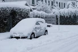 A car covered in snow is parked on a snow-covered street in front of a house. - Home Instead