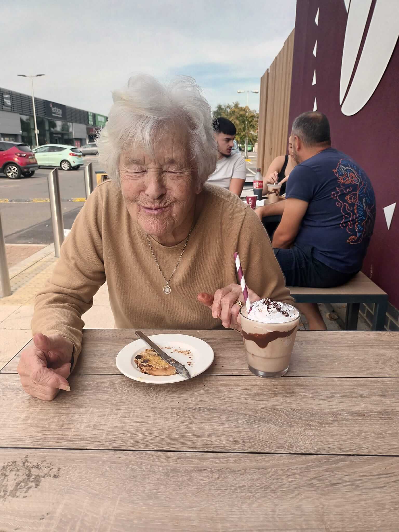 Elderly woman at a table with a dessert and milkshake, smiling with eyes closed in an outdoor seating area. - Home Instead