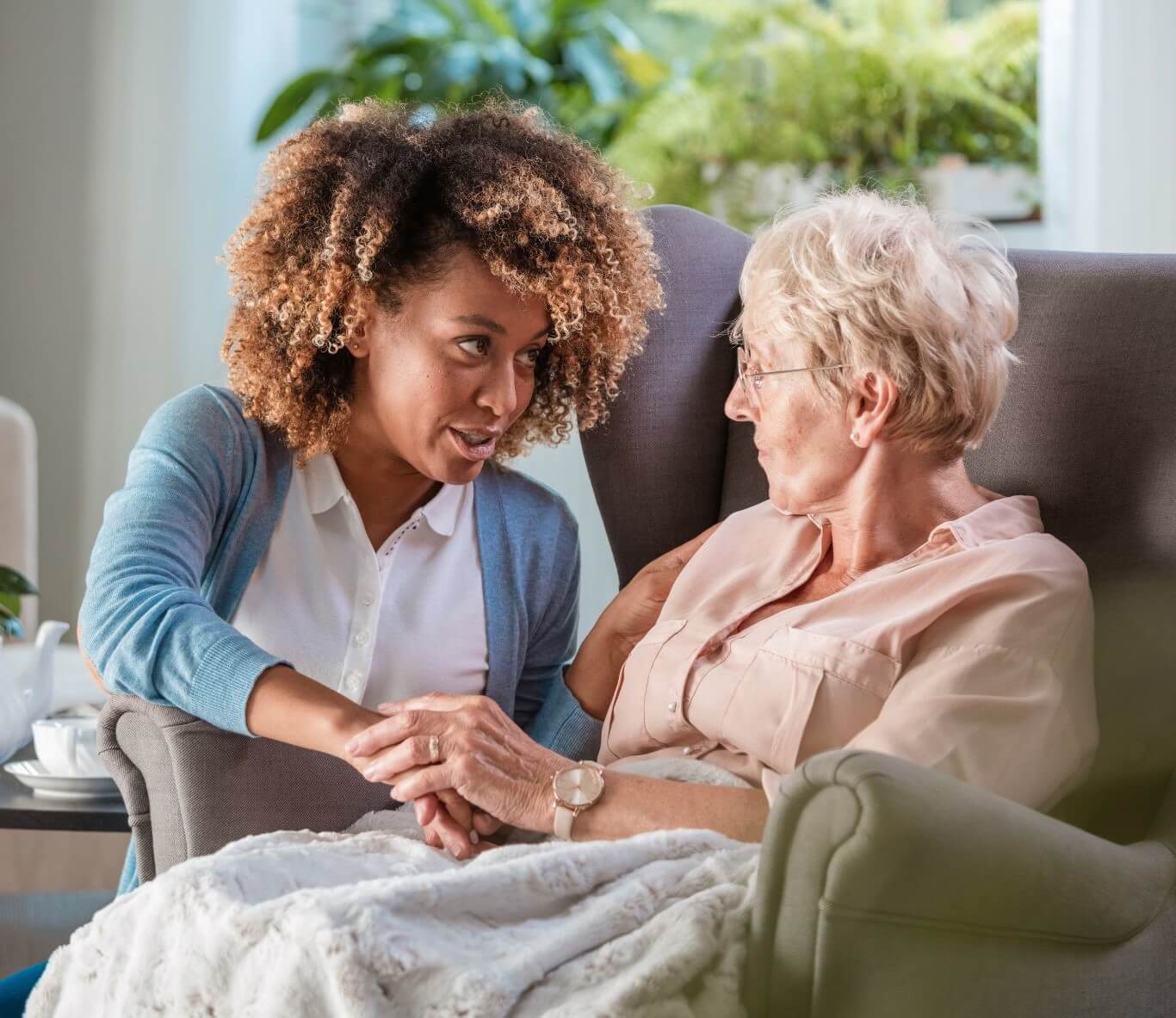A caregiver holds an elderly woman's hand and talks to her while sitting together in a cozy living room. - Home Instead
