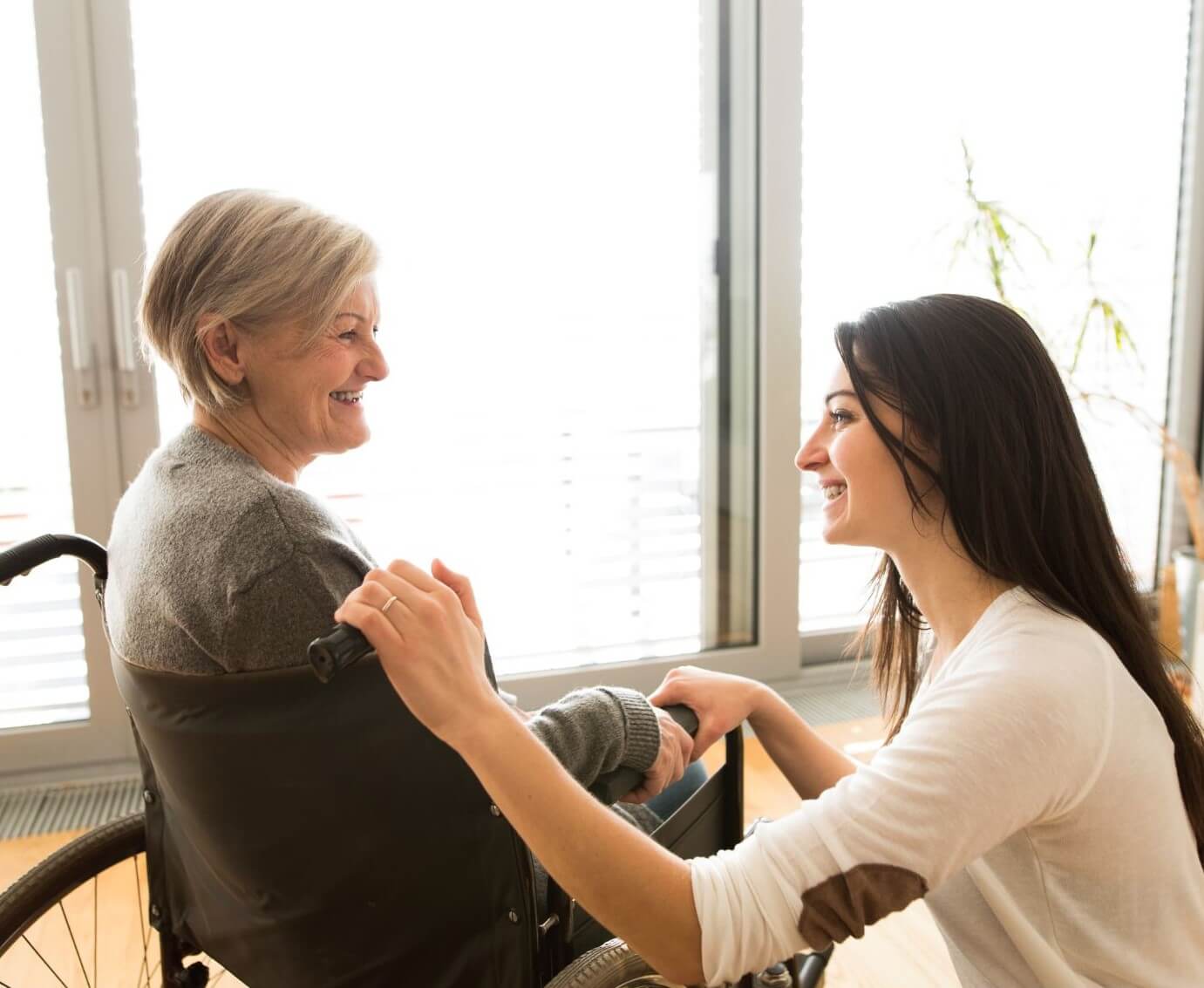 Young woman kneeling and holding hands with an elderly woman in a wheelchair, both smiling at each other. - Home Instead